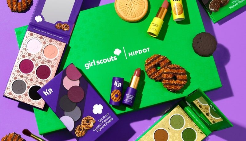 The HipDot X Girl Scouts Collection, featuring two eye shadow palettes and two lipsticks, on top of a green Girl Scouts box all on a purple background.