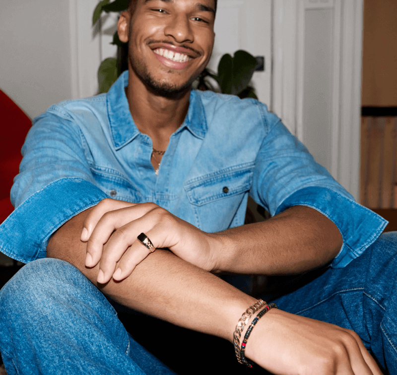 A man wearing all denim smiles while showing off a stack of bracelets and a ring on his left pinky finger