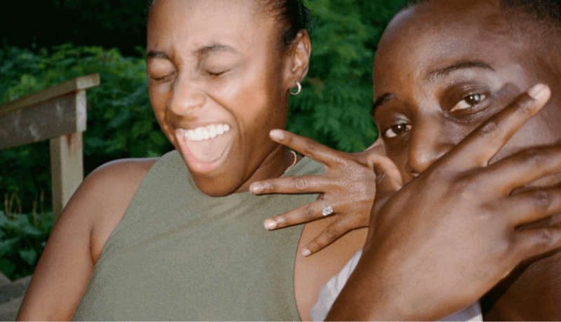 A woman looks excited as she shows off her engagement ring, while the man faces the camera with his hand over his mouth