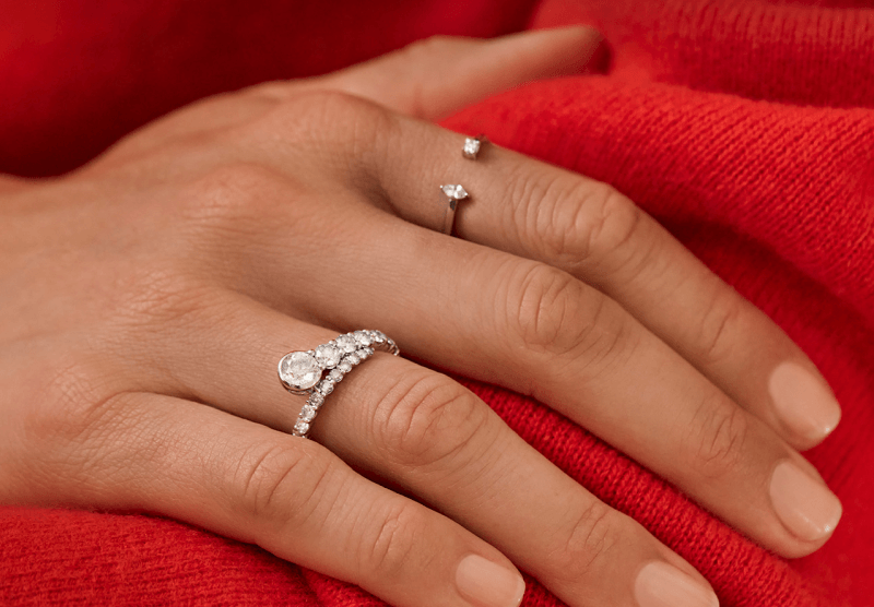 A woman wearing a red sweater rests her hand on her arm, showing off two stunning diamond rings