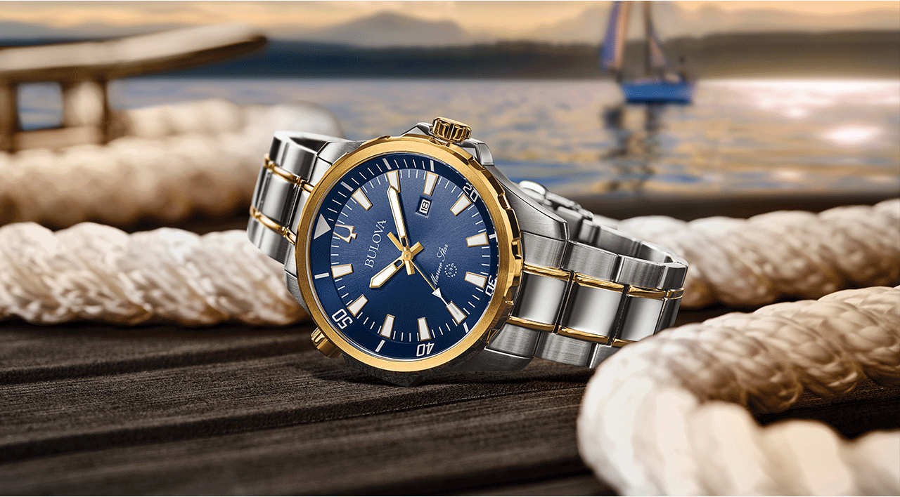 A Marine Star Series B in silver-tone stainless steel with gold-tone accents featuring a blue sun-ray dial with luminous hands sitting on a dock. Behind a sailboat on the water softly illuminated by a sun-setting lit sky.