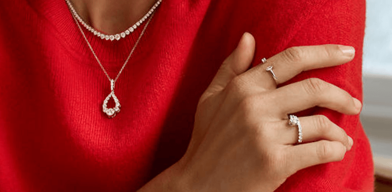 Cropped image of a woman in a red sweater where we see only her neck and hands, which show off stunning diamond necklace stacks and rings