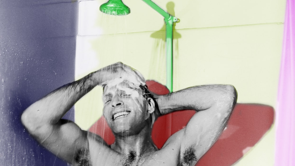 A black and white photo of a man showering and smiling as he washes his hair, with elements of the photo being hand colored in