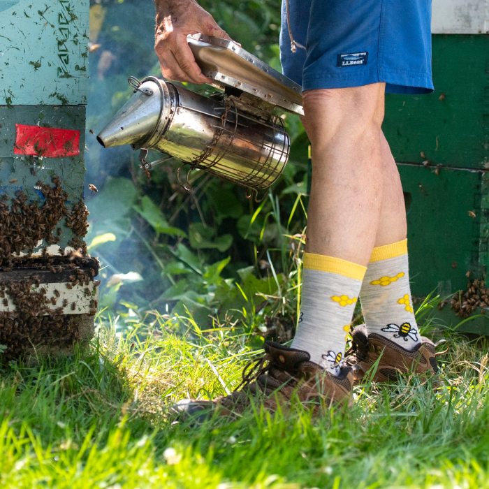 A beekeeper checking the hive wearing Darn Tough's new Knit to Give sock design, featuring bees and honeycomb