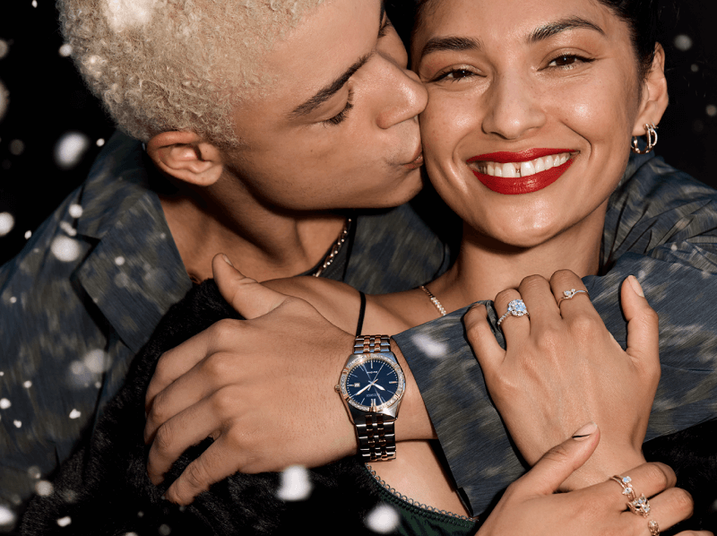 A couple embraces while showing off a stunning display of rings, necklaces, earrings, and a watch