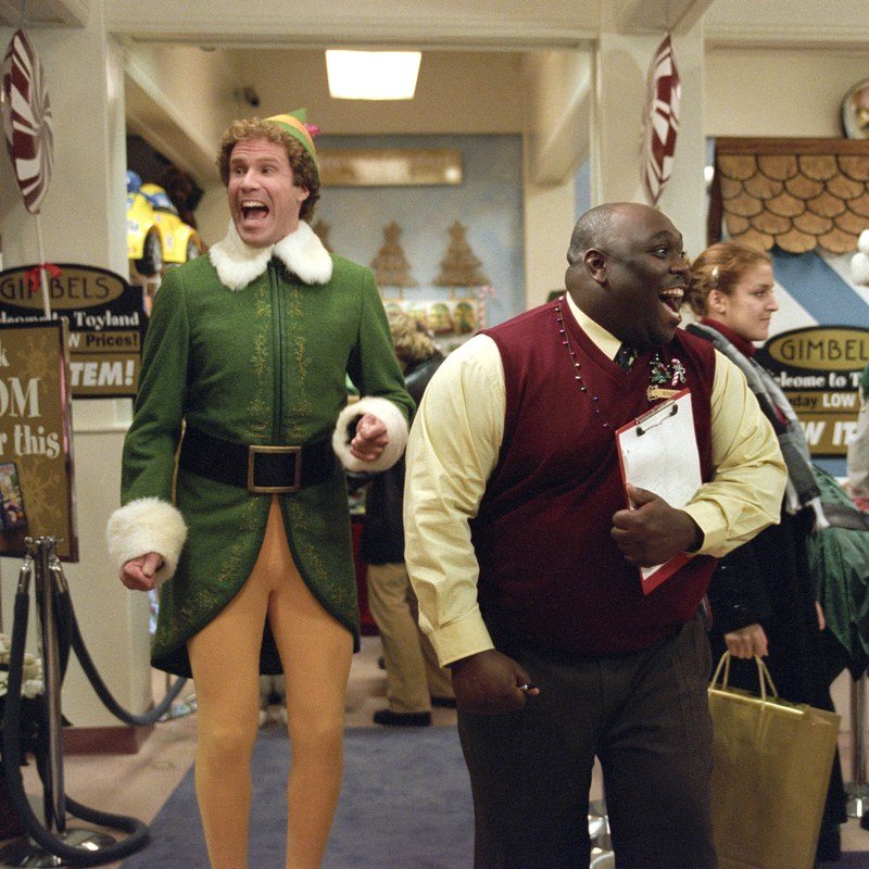 Will Ferrell in a green elf costume excited, jumping up and down in a shopping mall