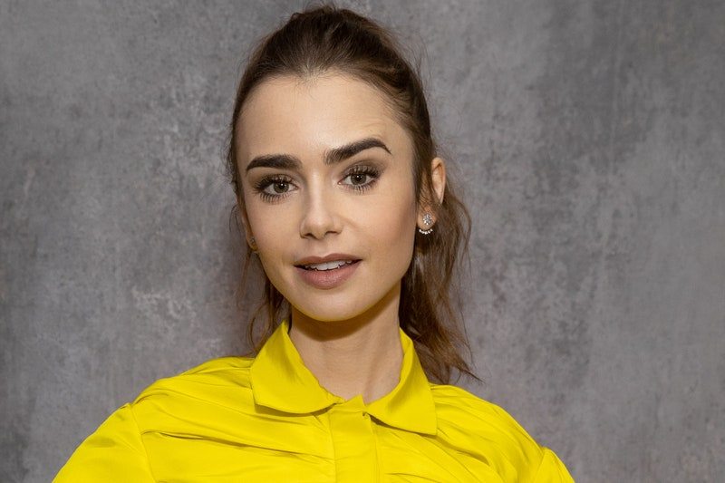 Lily Collins wears a ponytail and yellow blouse standing in front of a gray wall