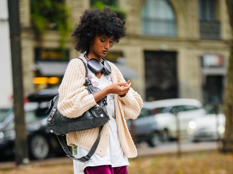 A guest wears a black headphones, a white t-shirt, a beige oversized wool cardigan, a black shiny leather nailed / studded City shoulder bag from Balenciaga (one of the it bags of 2025), purple shiny velvet large pants, black shiny leather platform soles boots , outside Dolce & Gabbana, during the Milan Fashion Week - Womenswear Spring/Summer 2023 on September 24, 2022 in Milan, Italy. 