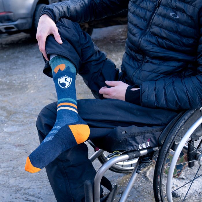 Person in a wheelchair holding up their foot to show the High Fives sock - a blue sock with a treeline design