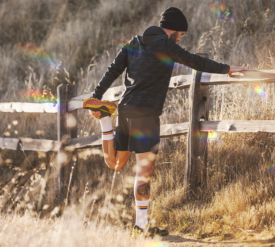 A runner in dark attire stretches his legs alongside a wooden fence in a beautiful sunlit field, striking in rainbow strapped socks.