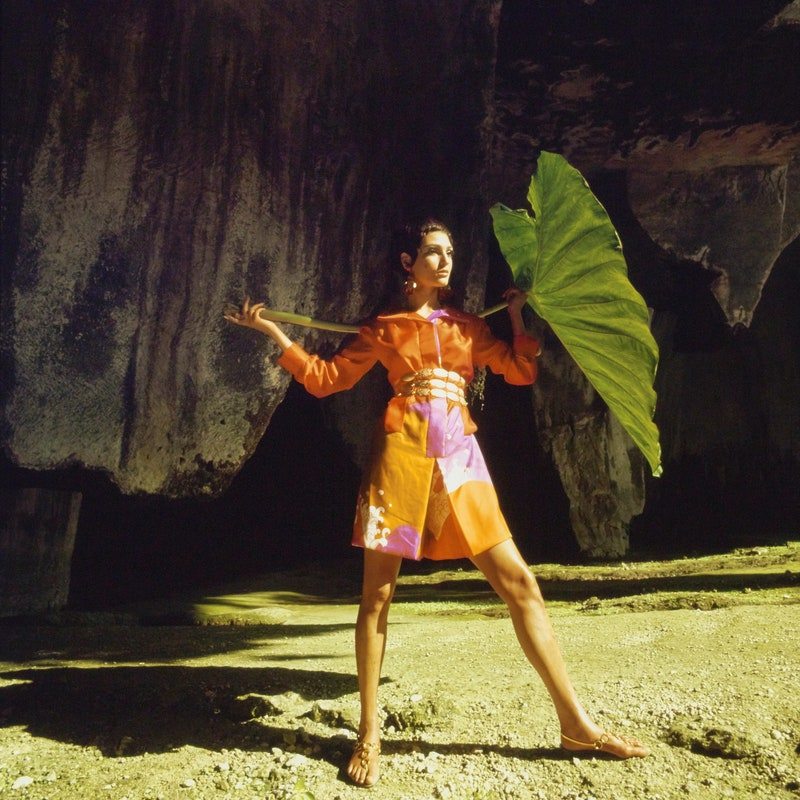 Model standing in the Orecchio di Dionisio, a high-vaulted groto hewn out of a quarry wall in Syracuse, Sicily, holding a huge palm leaf over her shoulder and wearing an orange and yellow patterned culotte-shirtdress by Hanae Mori, with gold hoop earrings by Robert Originals, a Napier gold chain belt and Boutique Marco gold sandals. *** Local Caption ***
