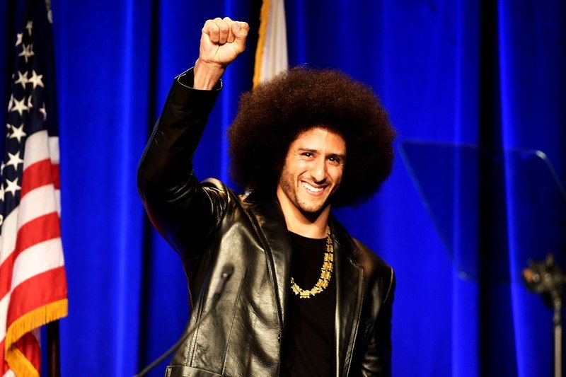 Colin Kaepernick raising his fist and smiling next to an American flag