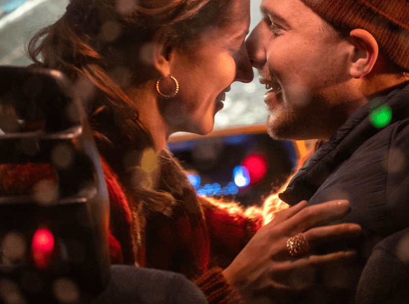 A couple lean in to kiss while our eye is drawn to the woman's earrings and ring