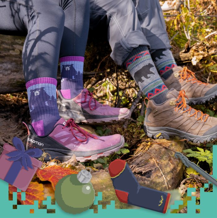 Shop Now: two hikers sit on a log in the woods, wearing bear-patterned Darn Tough socks