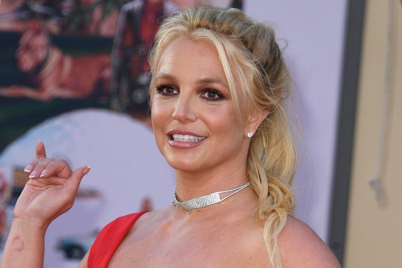 Britney Spears with blonde hair in ponytail wearing red dress and gold jewelry waving at event
