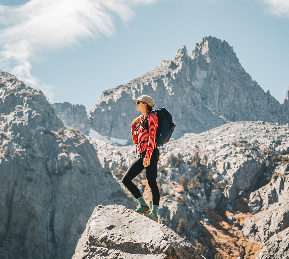 A hiker with a backpack stands atop a rocky mountain peak, taking it all in, rocking teal socks.