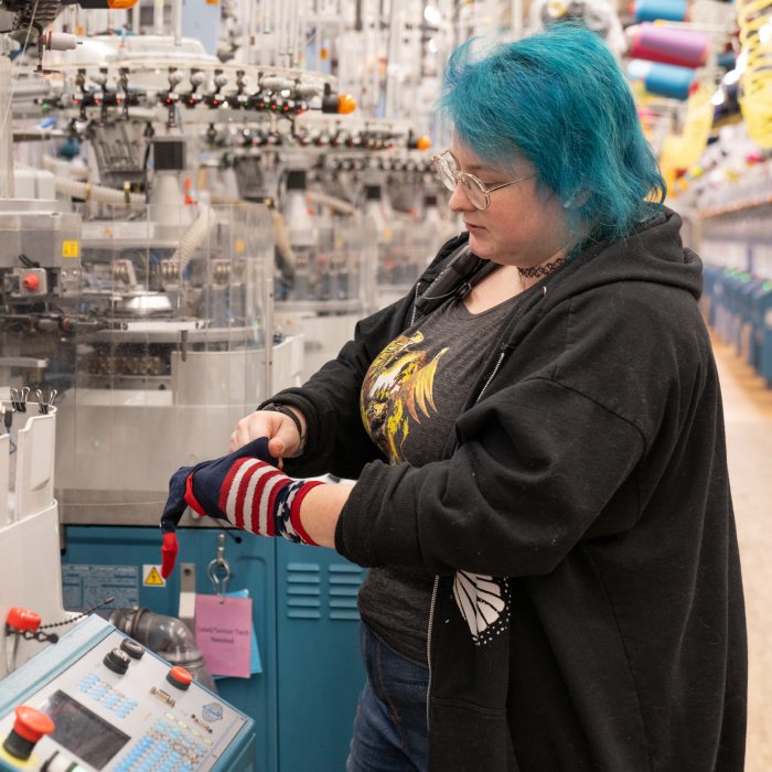 Our Mission - standing amongst rows of knitting machines, a Darn Tough employee inspects a red, white and blue sock