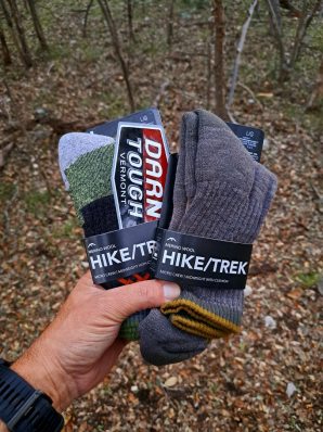 A reviewer in the woods holds two pairs of Darn Tough hike socks and a sticker