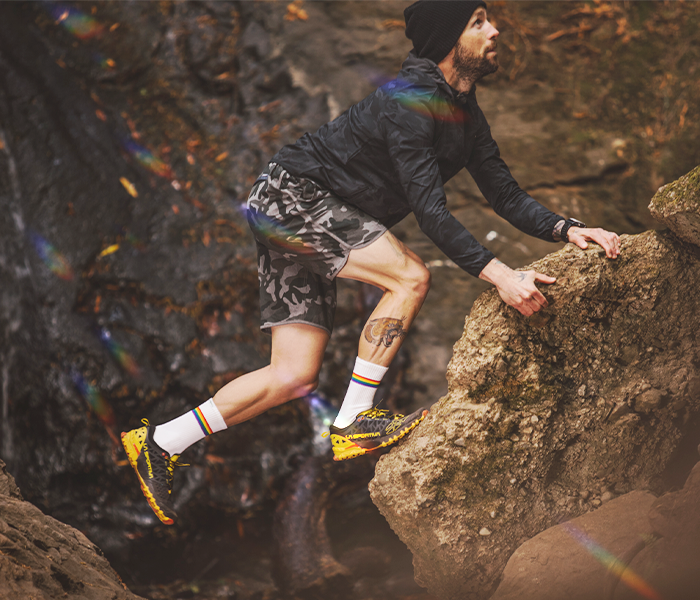 A guy with a leg tattoo leaps across rocks while slaying in our Prism socks.