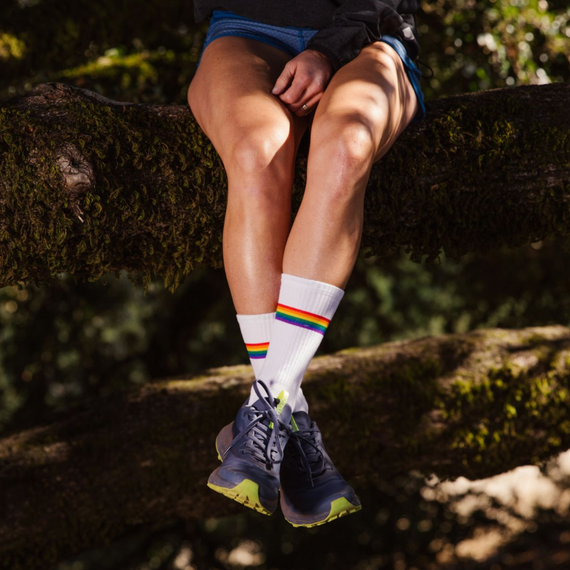 Someone sits on tree trunks while wearing sneakers, also tall white socks with a single rainbow band above the ankle. A kaleidoscopic effect.