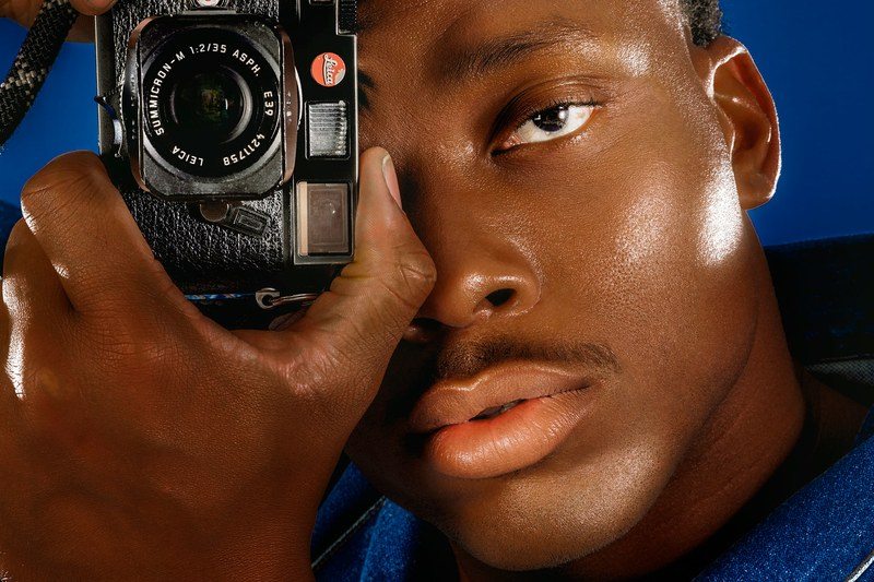 a tight shot of Romeo Okwara's face holding a camera to one eye wearing a denim jacket on a dark blue background