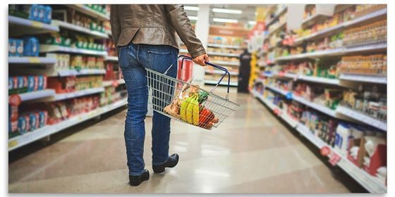 Back view of a person carrying a grocery basket in a supermarket aisle.