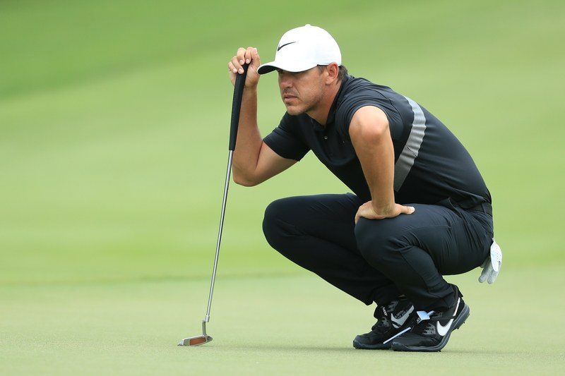 Brooks Koepka crouching down wearing a white baseball hat leaning on a golf club