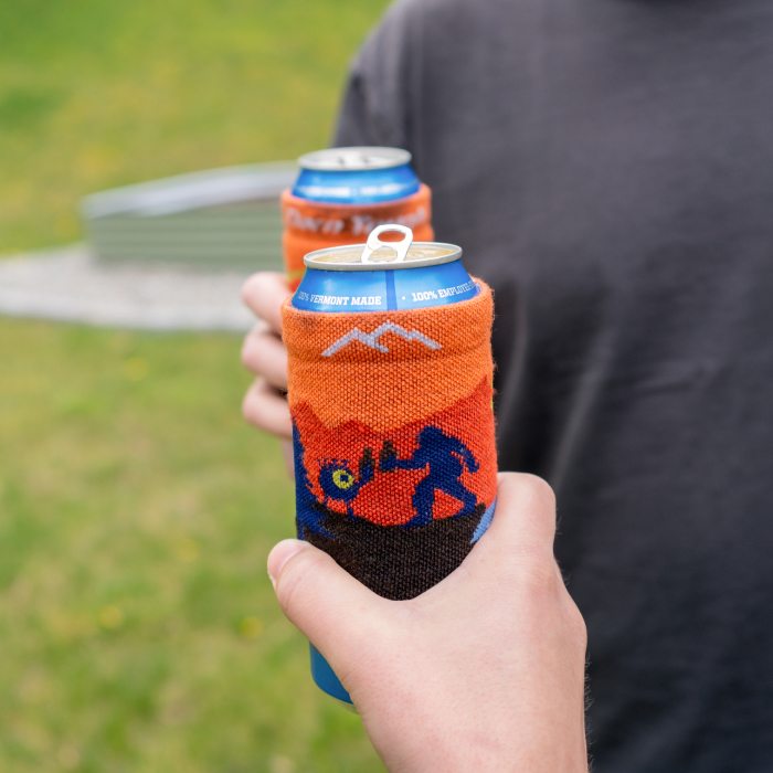 Shop now - people cheers with two cans, each inside an orange knit koozie featuring a mythical pattern