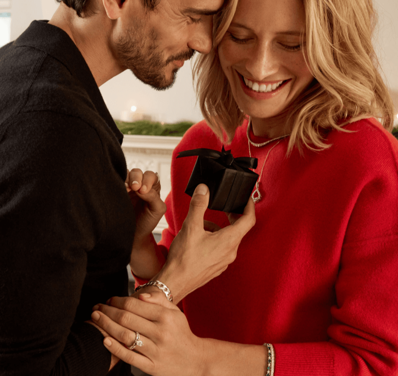 A couple huddle closely together, smiling over a small black gift box, and showing off delicate diamond jewelry