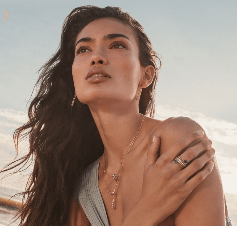 Woman with long hair at the beach wearing a grey strap dress, golden necklace, and jewelry, looking at someone.