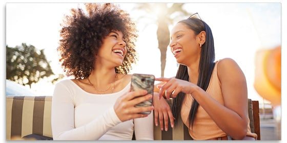 Two women sitting on a bench laughing and holding a phone.