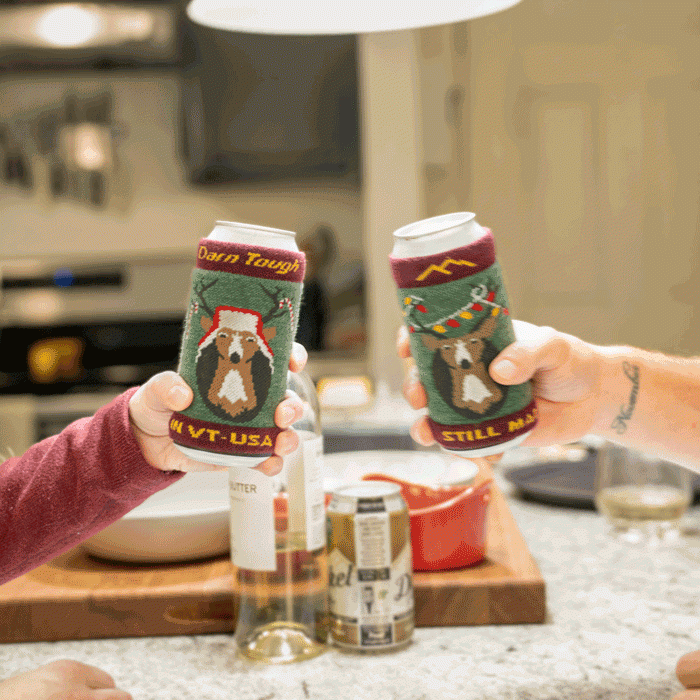 Two cans clinking, each wrapped in a Darn Tough knit koozie with reindeer holiday patterns.