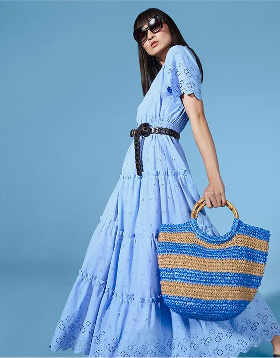 Woman wearing blue tiered maxi dress with broderie anglaise details, black belt, black sunglasses and blue-striped woven tote bag