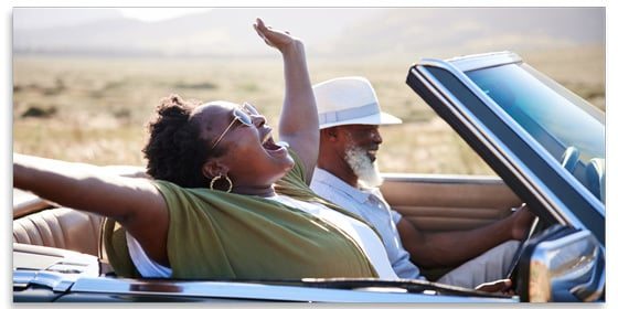 Man smiling and driving a convertible with the top down, and a smiling woman in the passenger seat with her hands in the air.