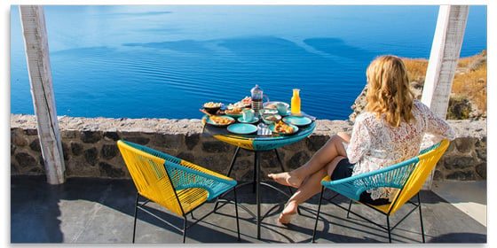 Woman sitting at a table laden with food on a cliff above the ocean on a sunny day.