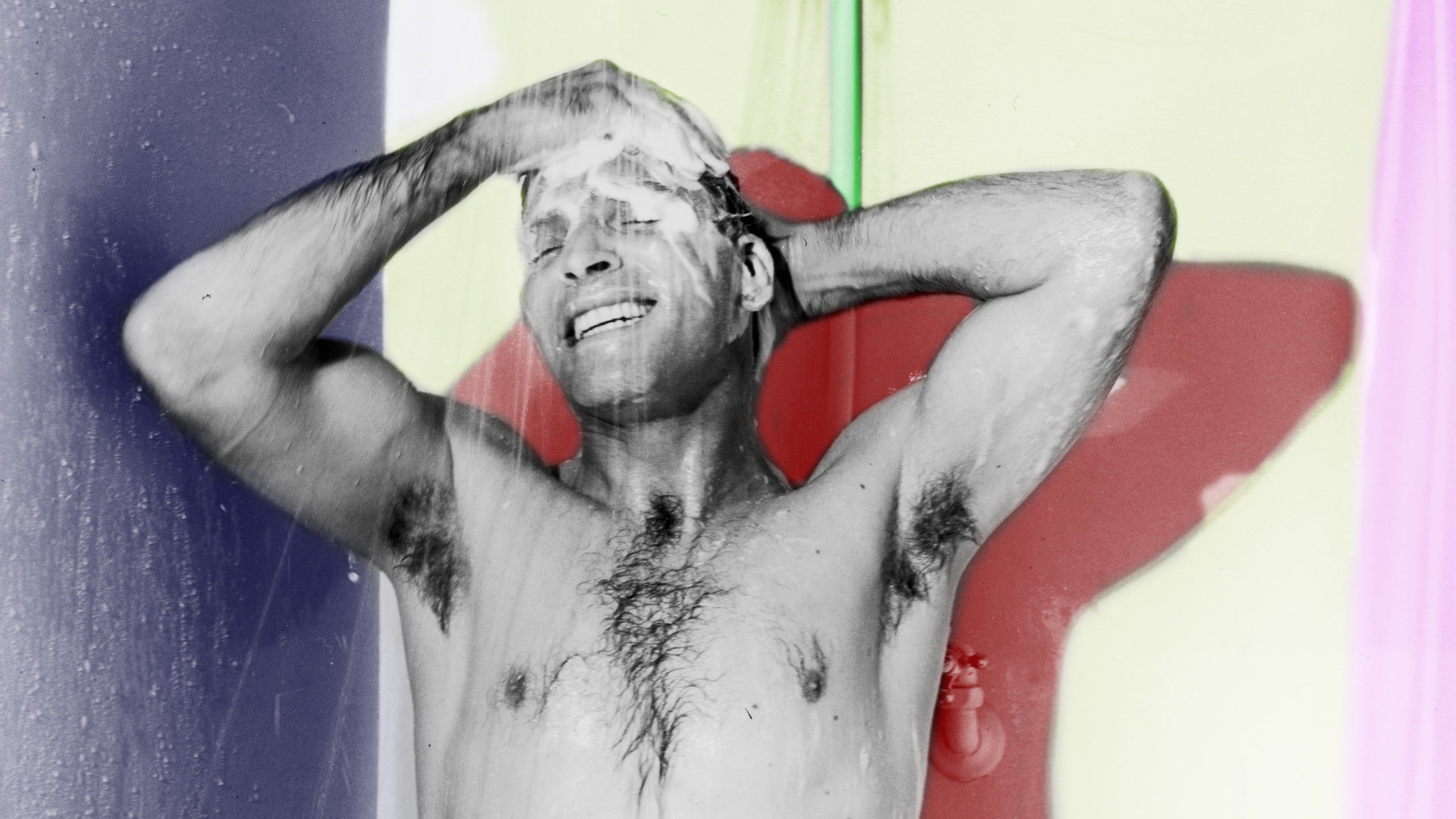 A black and white photo of a man showering and smiling as he washes his hair, with elements of the photo being hand colored in