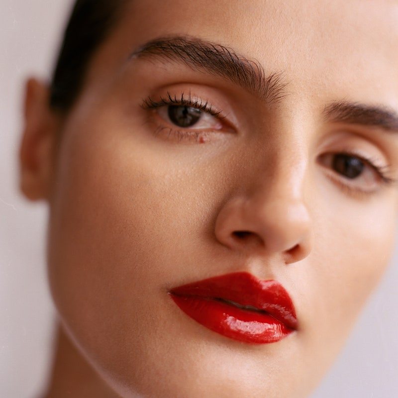 close-up shot of a model with medium skin tone, brown eyes, and thick eyebrows wearing a glossy, true red lip 