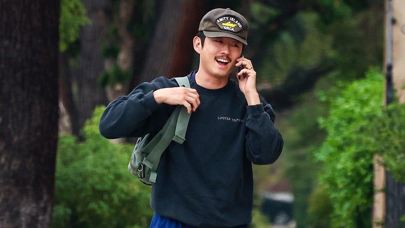 Steven Yeun on sidewalk talks on the phone while wearing a black sweatshirt, blue athletic shorts, army green shoulder bag, brown trucker hat, and dark green sneakers