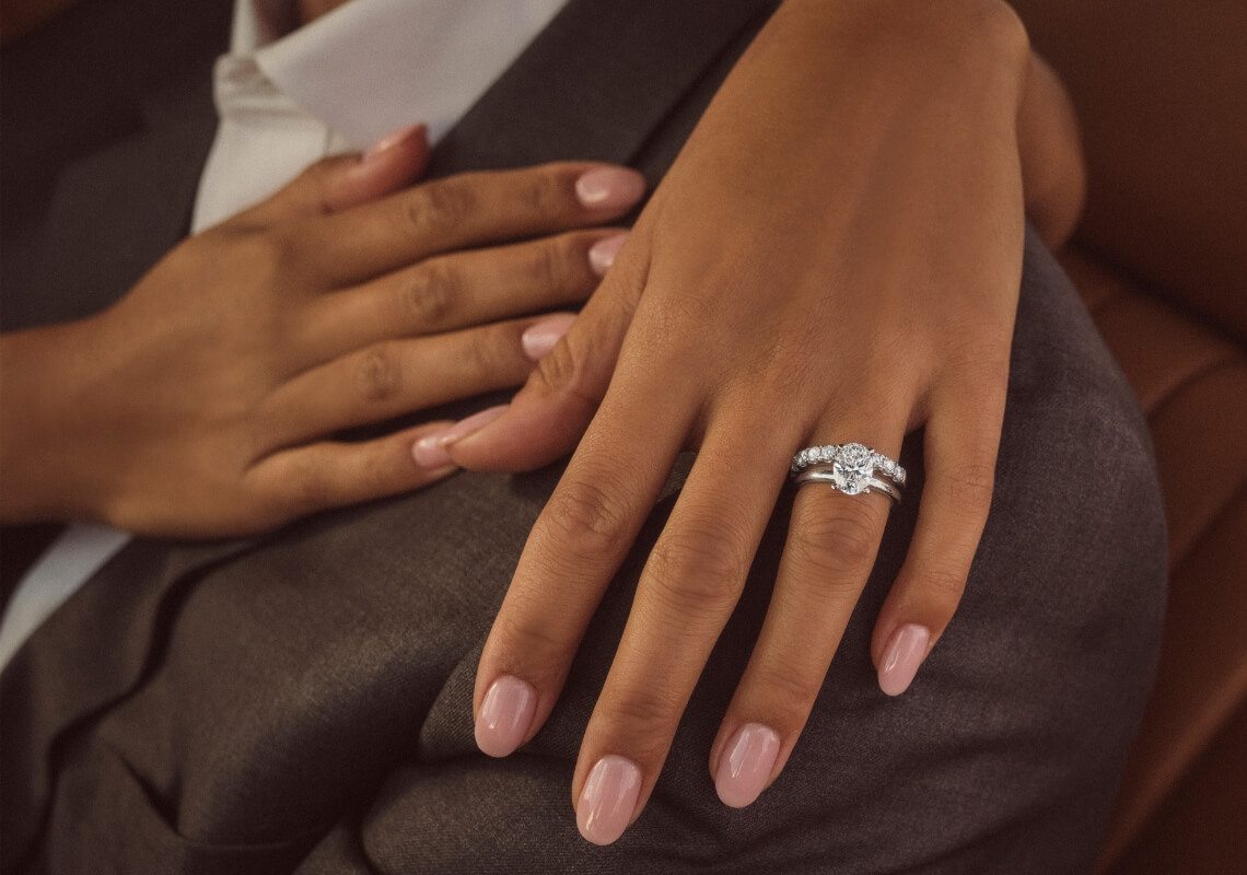 Close-up image of a woman's hand draped over man's shoulder with an oval-cut diamond engagement ring on her finger