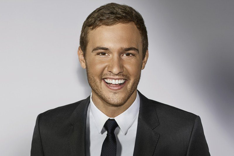 Peter Weber. the bachelor, wearing a suit smiling and holding a red rose on white background