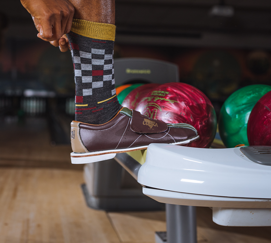 A guy at a bowling alley hoists his checkered Merino Wool socks up his calf, that big strike energy.