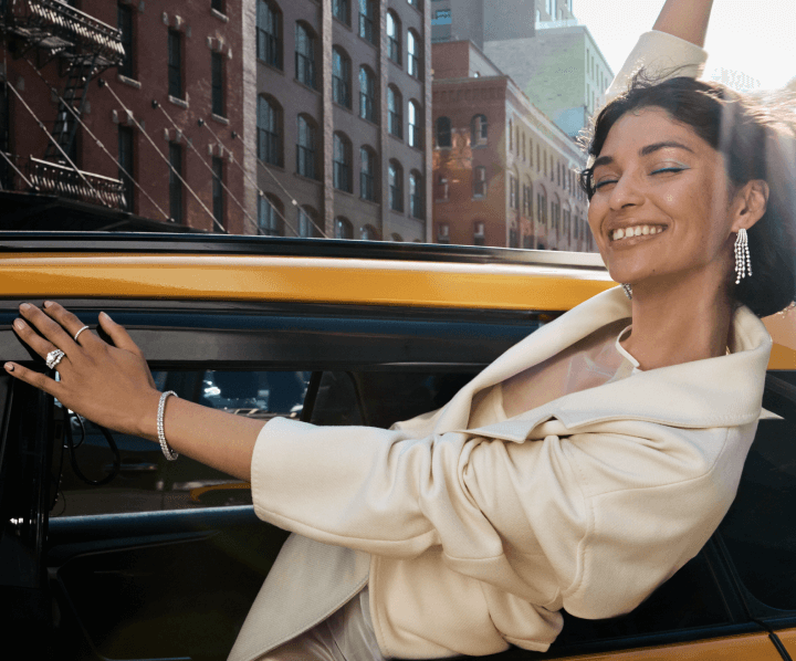 A smiling woman leans out of a car window. She is adorned with beautiful jewelry including diamond rings, necklaces, and earrings.