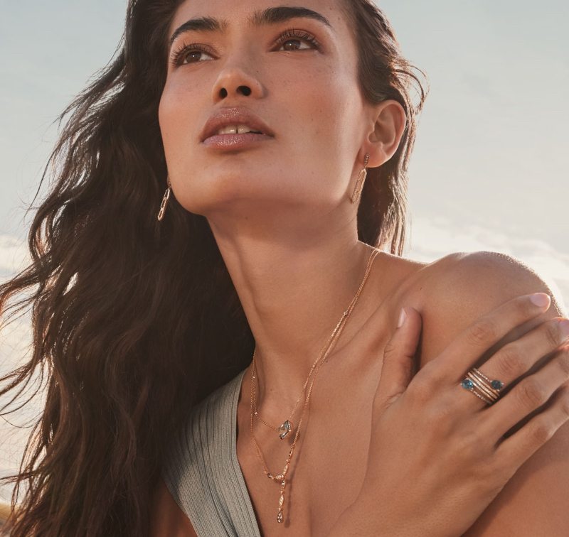 A woman with long, wavy hair wears layered gold necklaces and rings, looking at natural beauty of sky.