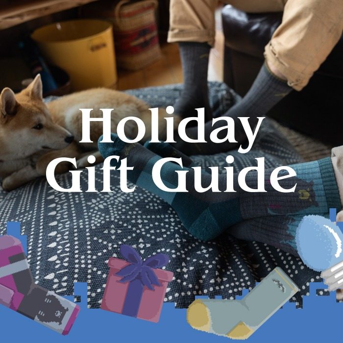 Shop the Holiday Gift Guide - a dog curled up on a blue ottoman, looking at the Darn Tough socks on their human's feet