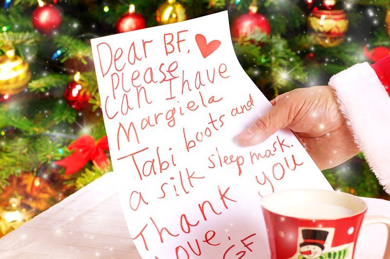 Santa's hand holding a Christmas list, in which a girlfriend asks her boyfriend for Margiela Tabi boots and a silk sleep mask
