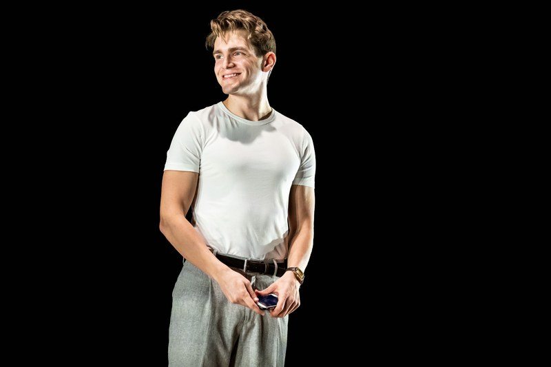 Andrew Burnap wearing a white t shirt tucked into belted slacks holding a cell phone in both hands smiling on a black stage