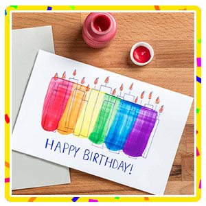 Rainbow colored candles drawn on a white page laying on a wood background with red paint