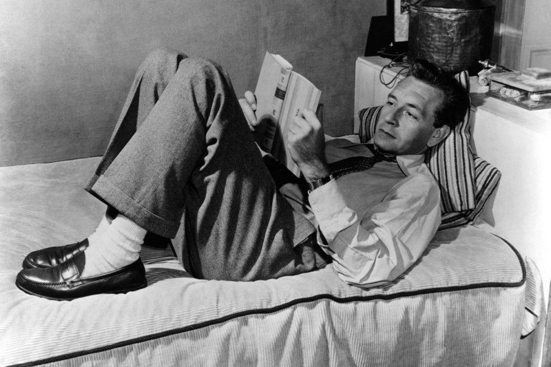 a black and white image of a man lying in bed wearing a shirt, slacks and loafers