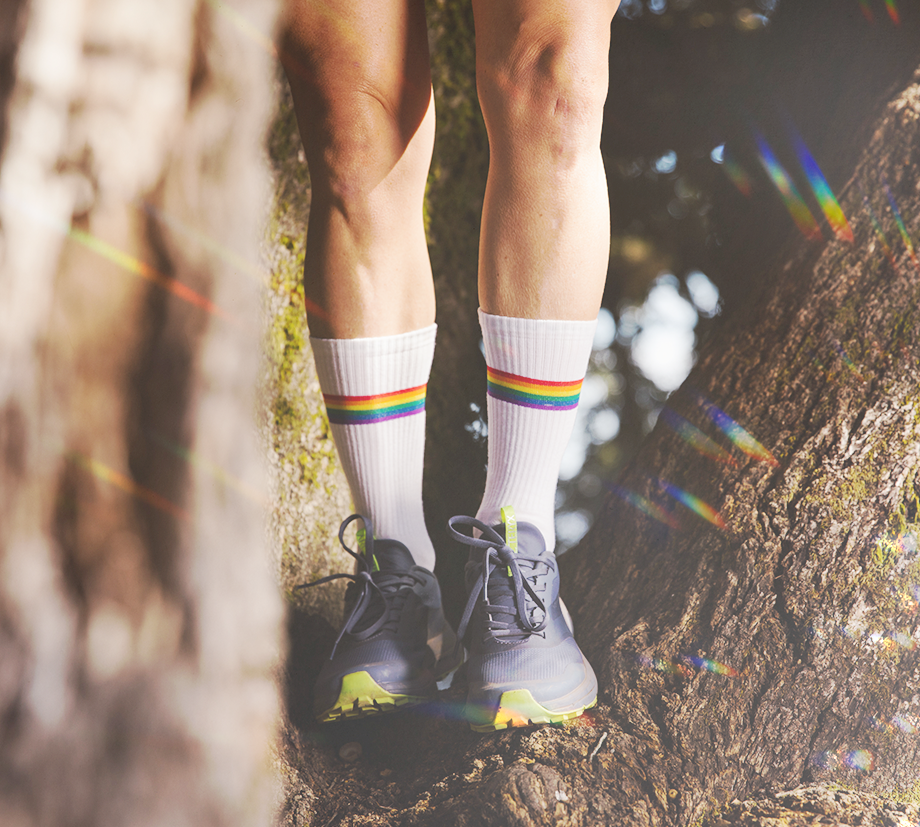 Someone stands on tree trunks while wearing sneakers, also tall white socks with a single rainbow band above the ankle. A kaleidoscopic effect.